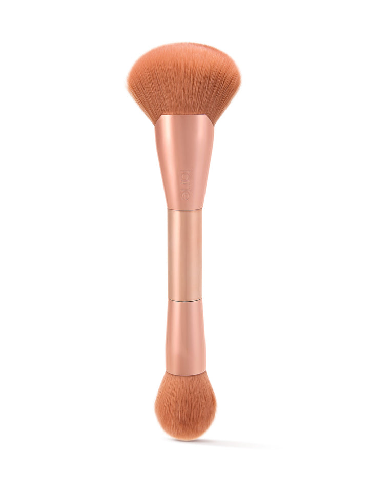 Dual-ended makeup brush with soft bristles; one end wide and fluffy, the other smaller and round. Held in a metallic handle, marked with Tarte, set against a white background.