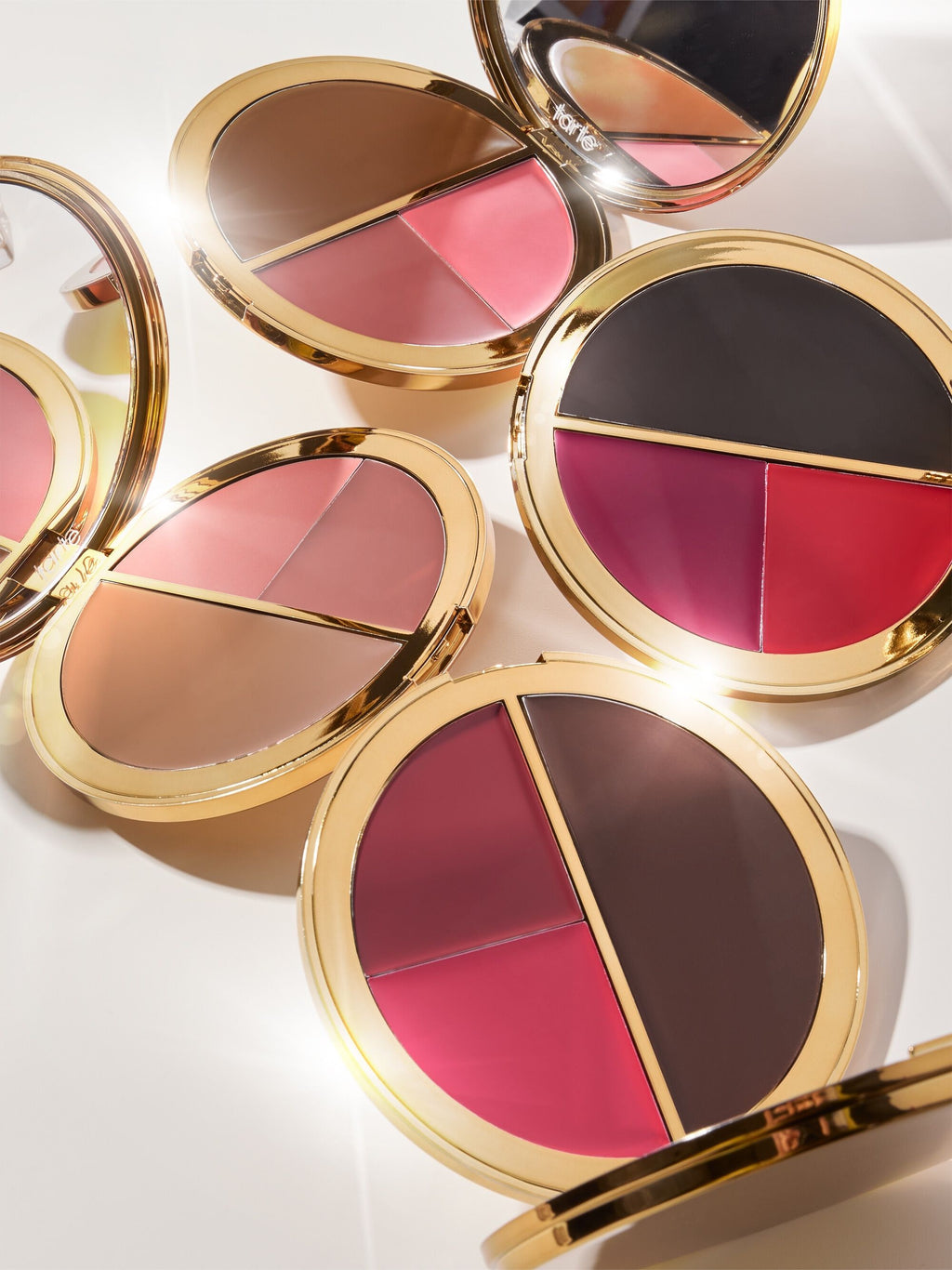 Compact makeup palettes with two sections of contrasting colors open to reveal shades of pink, brown, and red. Each palette features a reflective gold trim in a bright, well-lit setup. Text: tarte.