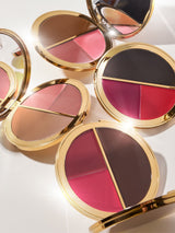 Compact makeup palettes with two sections of contrasting colors open to reveal shades of pink, brown, and red. Each palette features a reflective gold trim in a bright, well-lit setup. Text: tarte.
