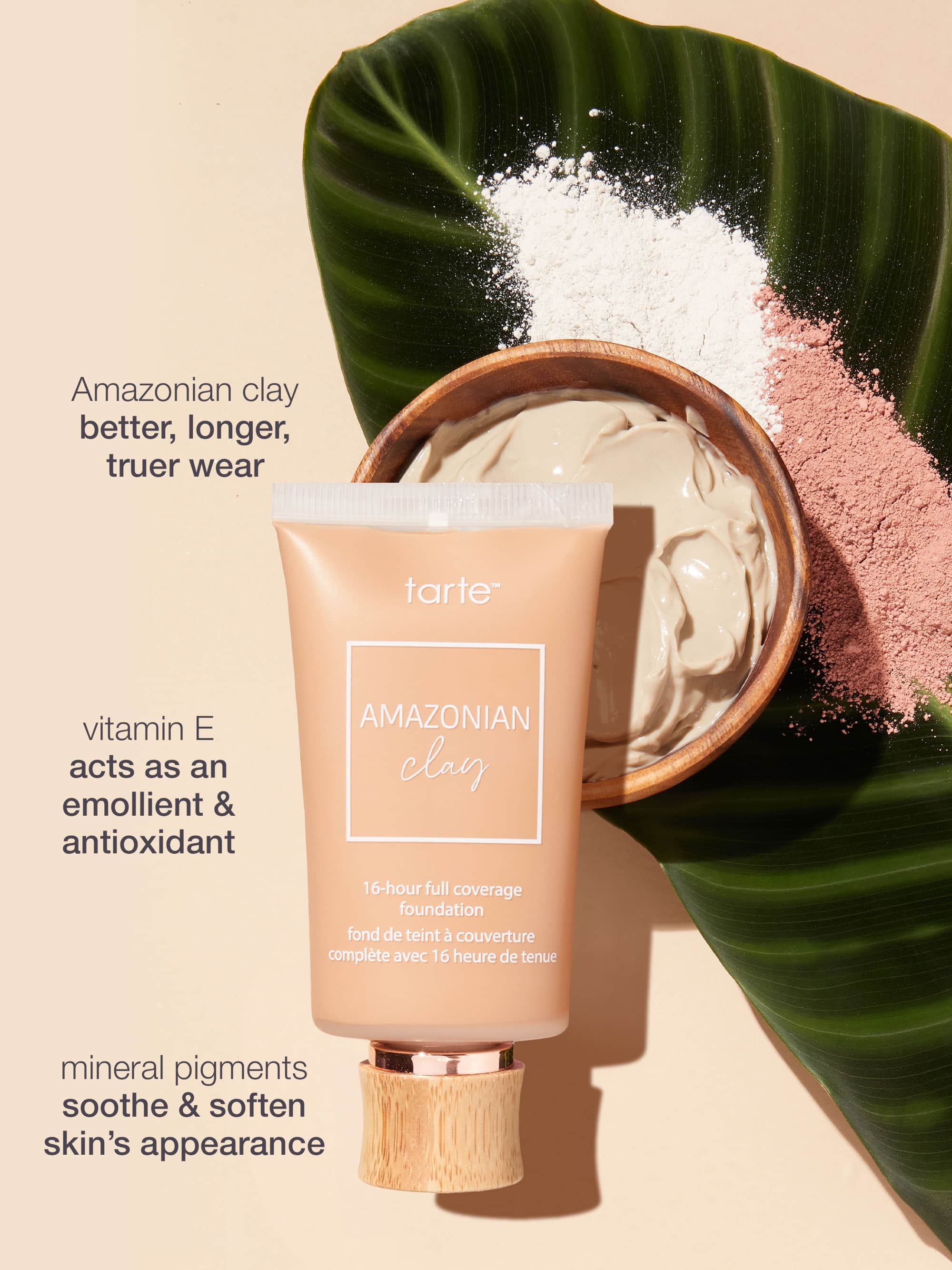A Tarte Cosmetics Amazonian clay foundation tube stands upright beside a wooden bowl of cream, atop a green leaf with pink and white powders. Text: Amazonian clay better, longer, truer wear. Vitamin E acts as an emollient & antioxidant. Mineral pigments soothe & soften skin’s appearance.