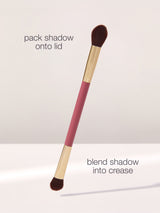 A dual-ended makeup brush with brown bristles, labeled for packing shadow onto the lid and blending into the crease, is displayed against a neutral background. Text: pack shadow onto lid, blend shadow into crease.