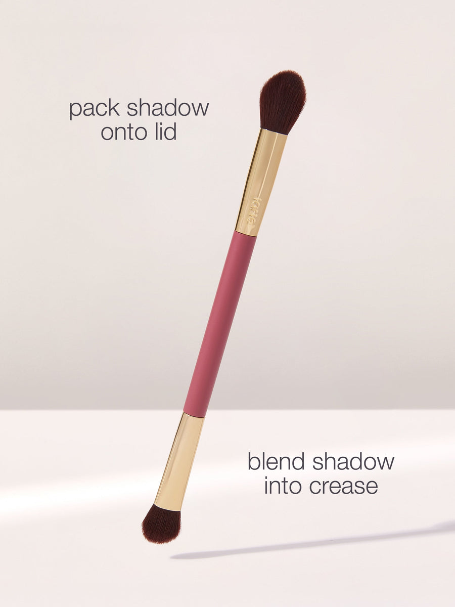 A dual-ended makeup brush with brown bristles, labeled for packing shadow onto the lid and blending into the crease, is displayed against a neutral background. Text: pack shadow onto lid, blend shadow into crease.