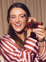 Woman applying makeup with a brush and compact mirror, wearing a striped shirt.