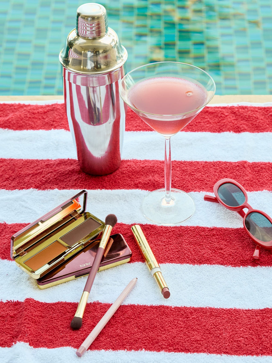 Cocktail shaker, pink cocktail, sunglasses, makeup set on a red and white striped towel by a pool.