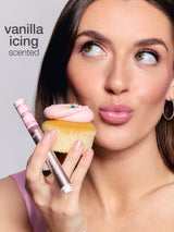 Tarte Cosmetics lip products, including Plumping Lip Oil, Melt Mask, Juicy Lip, Lip Plump, and Lip Vinyl, stand upright in front of stacked donuts with pink icing. Text: vanilla icing scent.