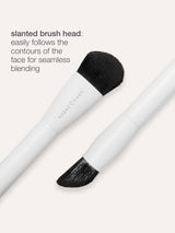 Makeup brush with a slanted head designed for seamless blending, lying against a plain background. Text reads: slanted brush head: easily follows the contours of the face for seamless blending.