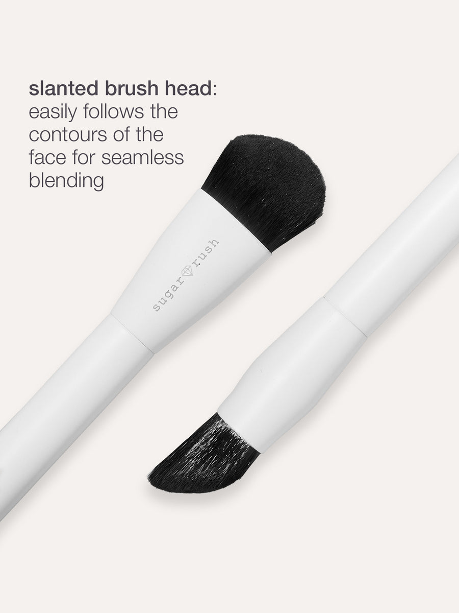 Makeup brush with a slanted head designed for seamless blending, lying against a plain background. Text reads: slanted brush head: easily follows the contours of the face for seamless blending.