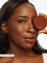 A woman holds a Tarte Cosmetics compact near her eye, displaying smooth skin and glossy lips, against a plain white background. Text at the bottom reads Grace in confident.