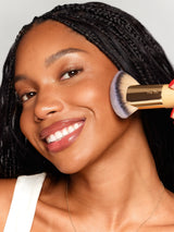 Woman applying makeup with a brush, wearing a white top against a neutral background