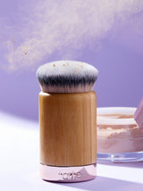 A Tarte Cosmetics brush with dense bristles disperses fine powder into the air, set against a lavender background. A jar of powder is partially visible in the background.