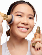 Woman applying makeup with a brush and bottle, smiling against a plain background