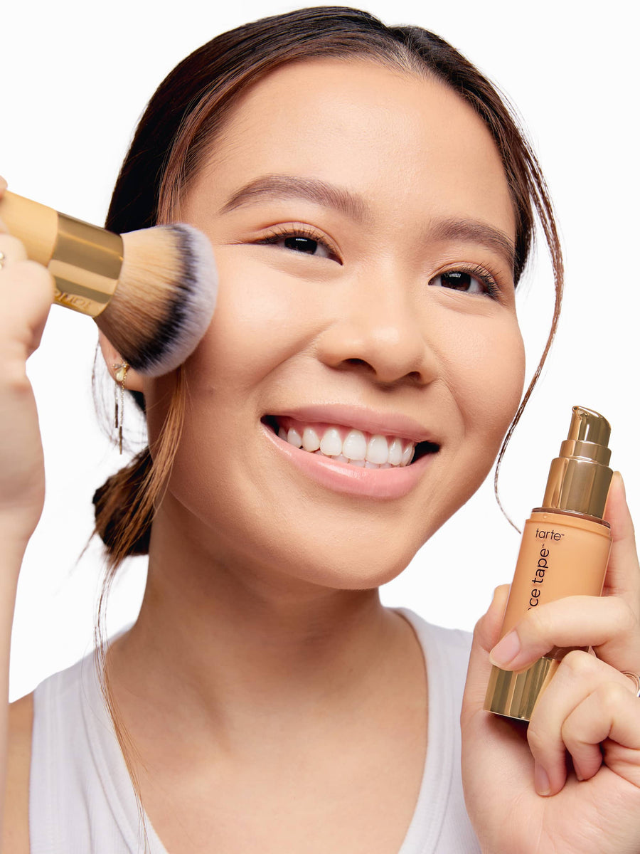 Woman applying makeup with a brush and bottle, smiling against a plain background