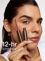 A person holds Tarte Cosmetics' brow product to their eyebrow, showcasing its application. Text reads, 12-hr longwear, highlighting the product's durability, against a plain background.