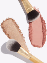 Makeup brushes spread shimmering beige and rosy pink powders on a clean white background. Each brush has a gold and wooden handle embossed with tarte.
