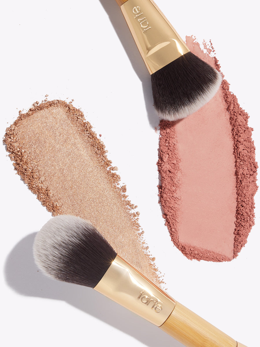Makeup brushes spread shimmering beige and rosy pink powders on a clean white background. Each brush has a gold and wooden handle embossed with tarte.