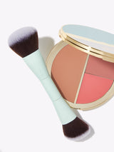A makeup brush with a green handle lies beside a round, open compact containing three color sections. The background is white and minimal. tarte is visible on the brush handle.