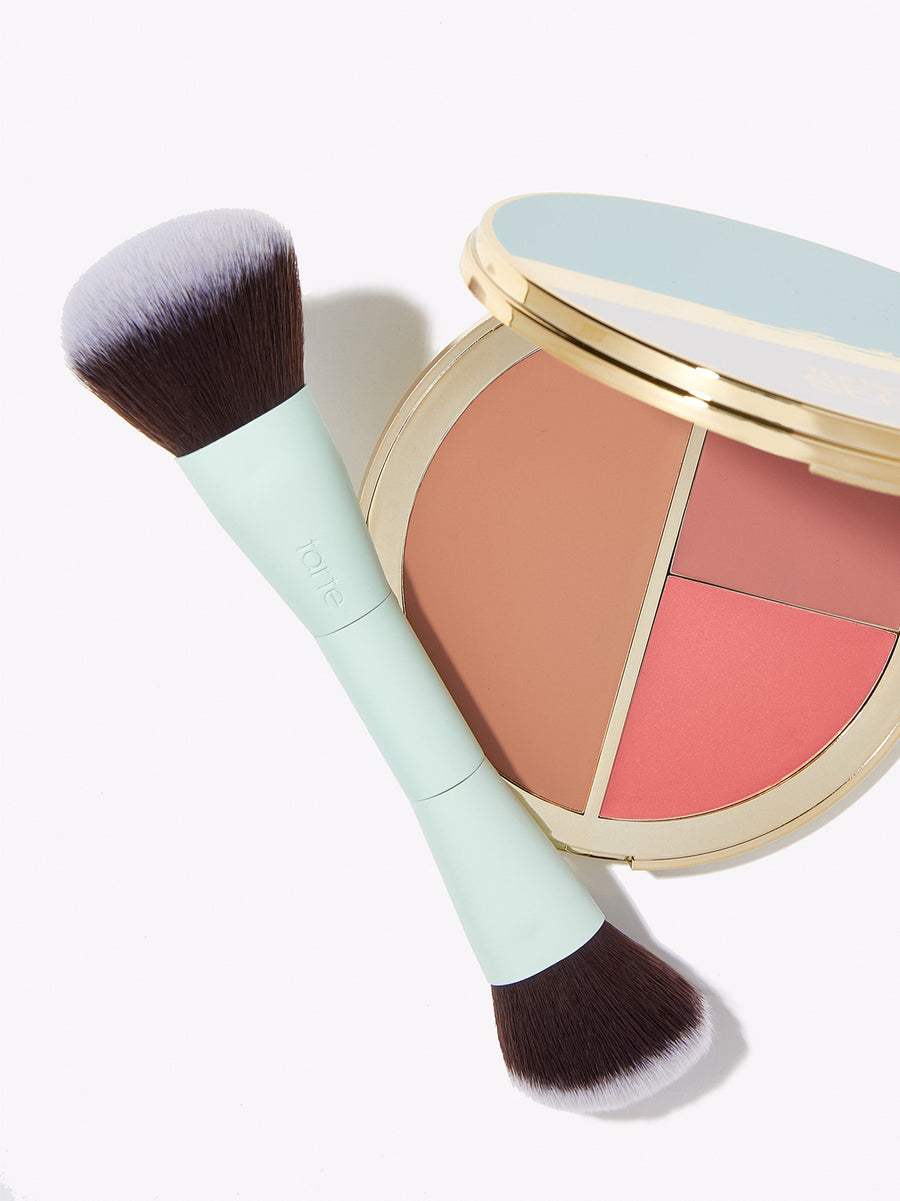 A makeup brush with a green handle lies beside a round, open compact containing three color sections. The background is white and minimal. tarte is visible on the brush handle.