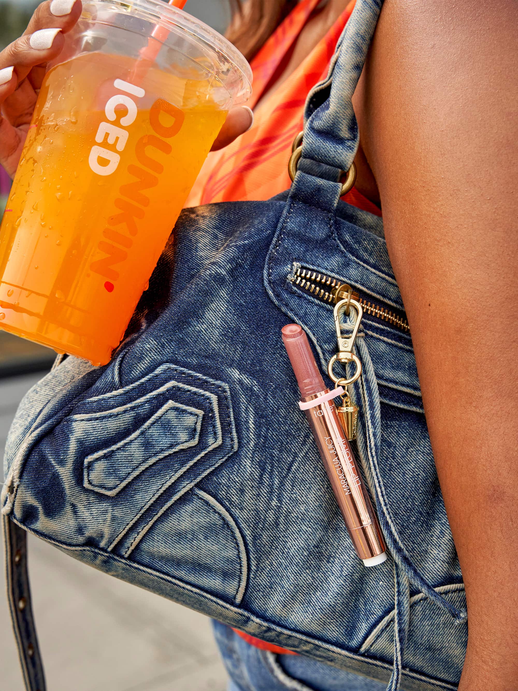 Person holding a Dunkin' iced coffee cup with a denim bag and pink lip gloss.