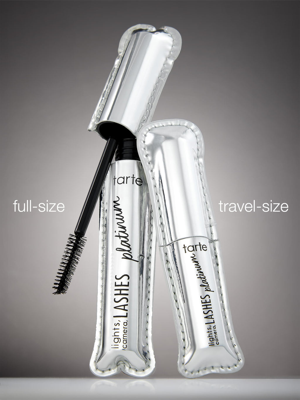 A shiny silver mascara tube labeled tarte lights, camera, LASHES platinum with a brush applicator exposed. It's accompanied by a travel-sized version, both on a reflective surface. Text: full-size, travel-size.