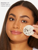 Woman holding a white round makeup pad with black specks, against a neutral background.