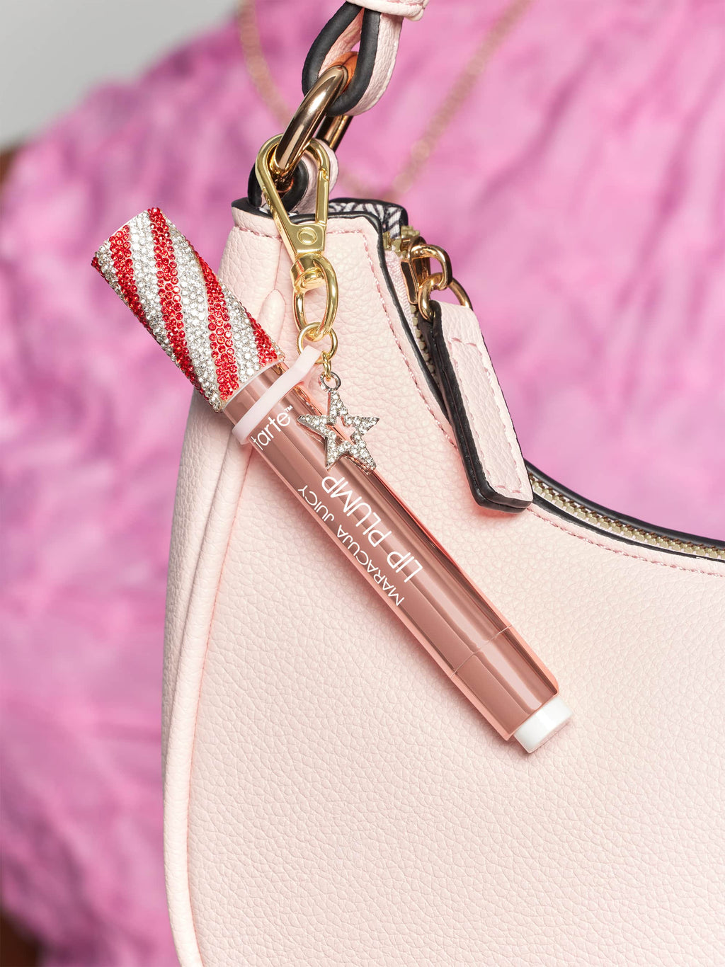 Lip gloss attached to a pink handbag. The gloss has red and white striped cap with rhinestones and a charm. Text reads: Tarte maracuja juicy lip plump. Background is soft pink fabric.