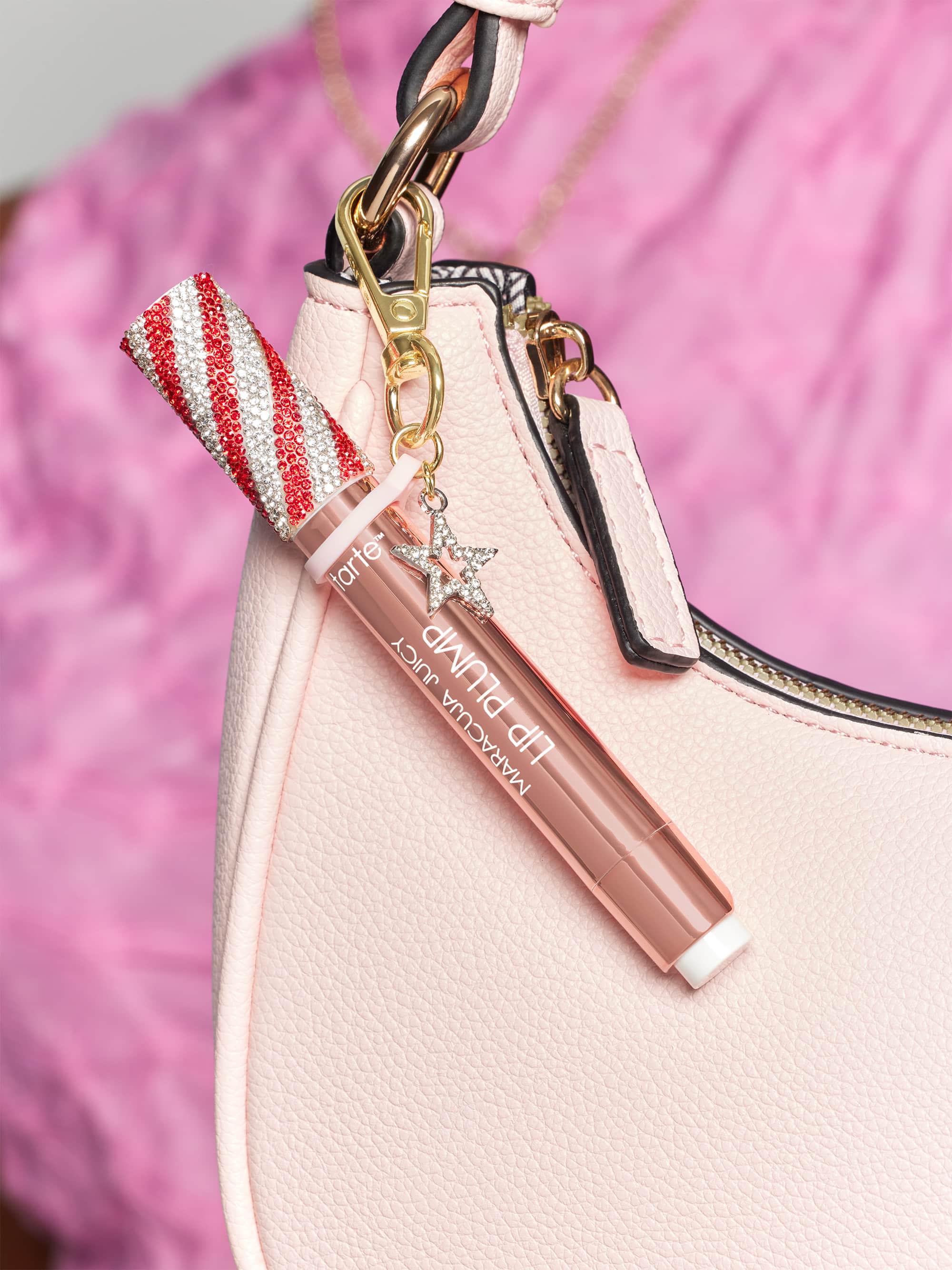 Lip gloss attached to a pink handbag. The gloss has red and white striped cap with rhinestones and a charm. Text reads: Tarte maracuja juicy lip plump. Background is soft pink fabric.