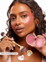 Open Tarte compact and brush held near face as model applies pink cream and shimmery blush; close-up against a plain white background. Text on image: Remmi in strawberry bliss