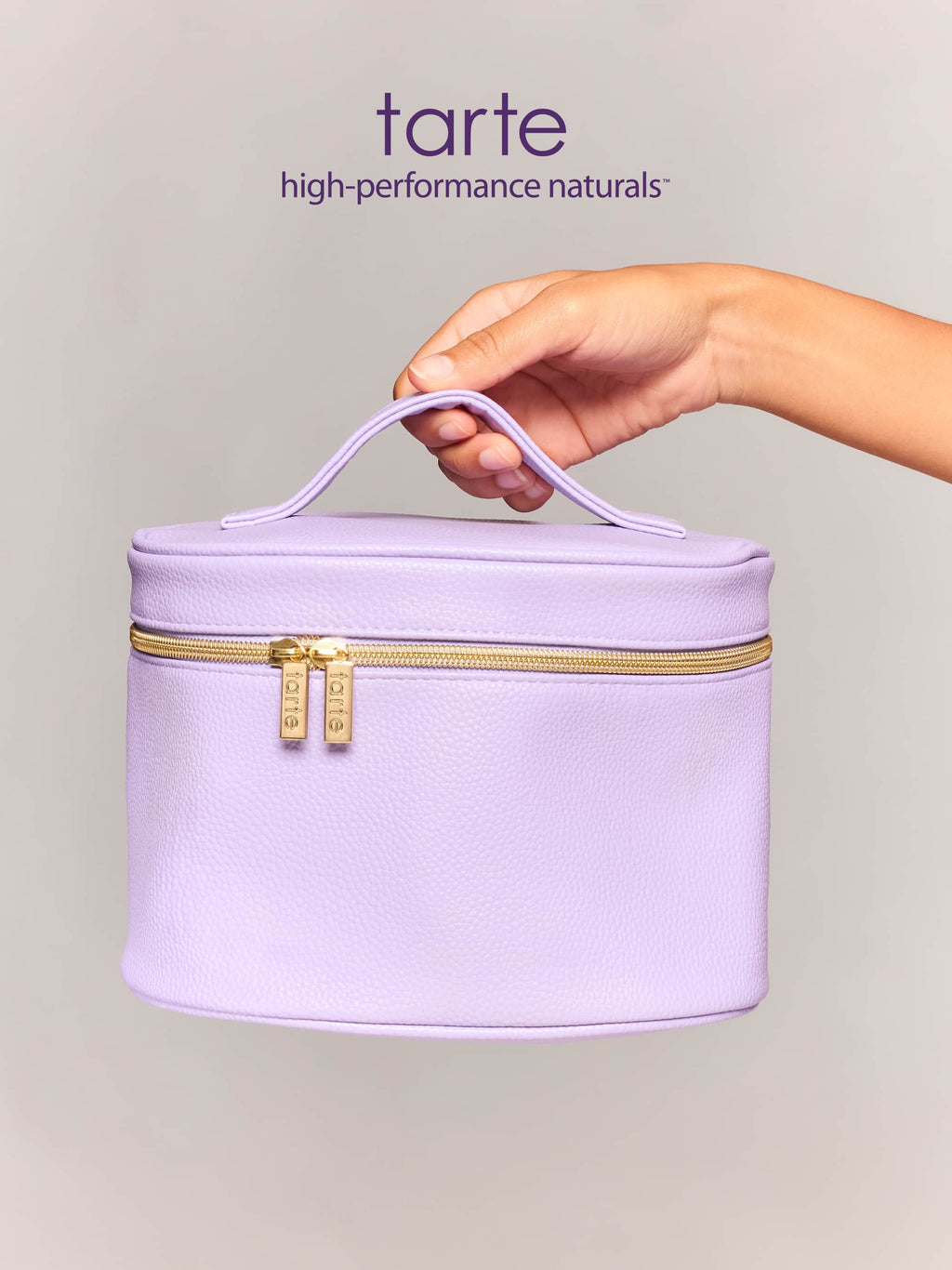 A lavender makeup bag with a handle is held by a hand, showcasing a zippered closure with two gold zippers labeled Tarte, against a plain, light gray background.