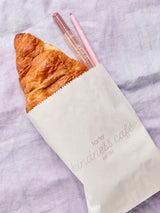 A croissant and two lip products from Tarte Cosmetics peek out of a paper bag labeled tarte kindness café est. '99, resting on a light purple fabric background.