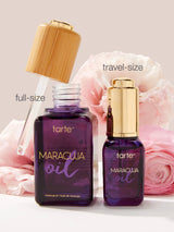 Two bottles of tarte Maracuja oil, one full-size and one travel-size, with pink flowers in the background.