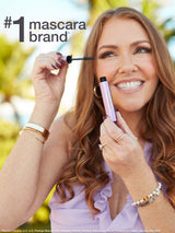 Woman holding a Tarte mascara tube and wand, applying mascara to her upper lashes while smiling outdoors with blurred palm trees in the background. Text: #1 mascara brand* *Source: Circana, LLC, U.S. Prestige Beauty Total Measured Market, Makeup Mascara by Brand, Dollar & Unit Sales, January–May 2025