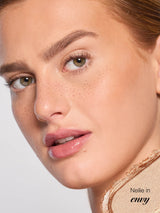 Close-up of a young woman's face, gazing slightly right with glossy lips, natural-looking freckles, defined brows and dewy skin; soft studio lighting and a textured gold cosmetic on her shoulder. Text: Neilie in envy