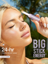 A lavender cosmetic stick is held near a person's closed eyelid; sunlit outdoor background with blurred palm trees. Text on image: 24-hr crease-proof wear; Lex in 20B; BIG STICK ENERGY