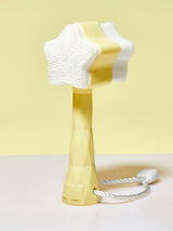 A star-shaped, dual-sided facial brush with soft bristles and textured nubs. It stands on a yellow handle engraved with a diamond and letters, set against a pastel background.