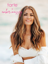 A woman with long, wavy hair smiles slightly, wearing a white top and open shirt. She stands outdoors near a blurred beach. Text reads: tarte x amber massey in pink.