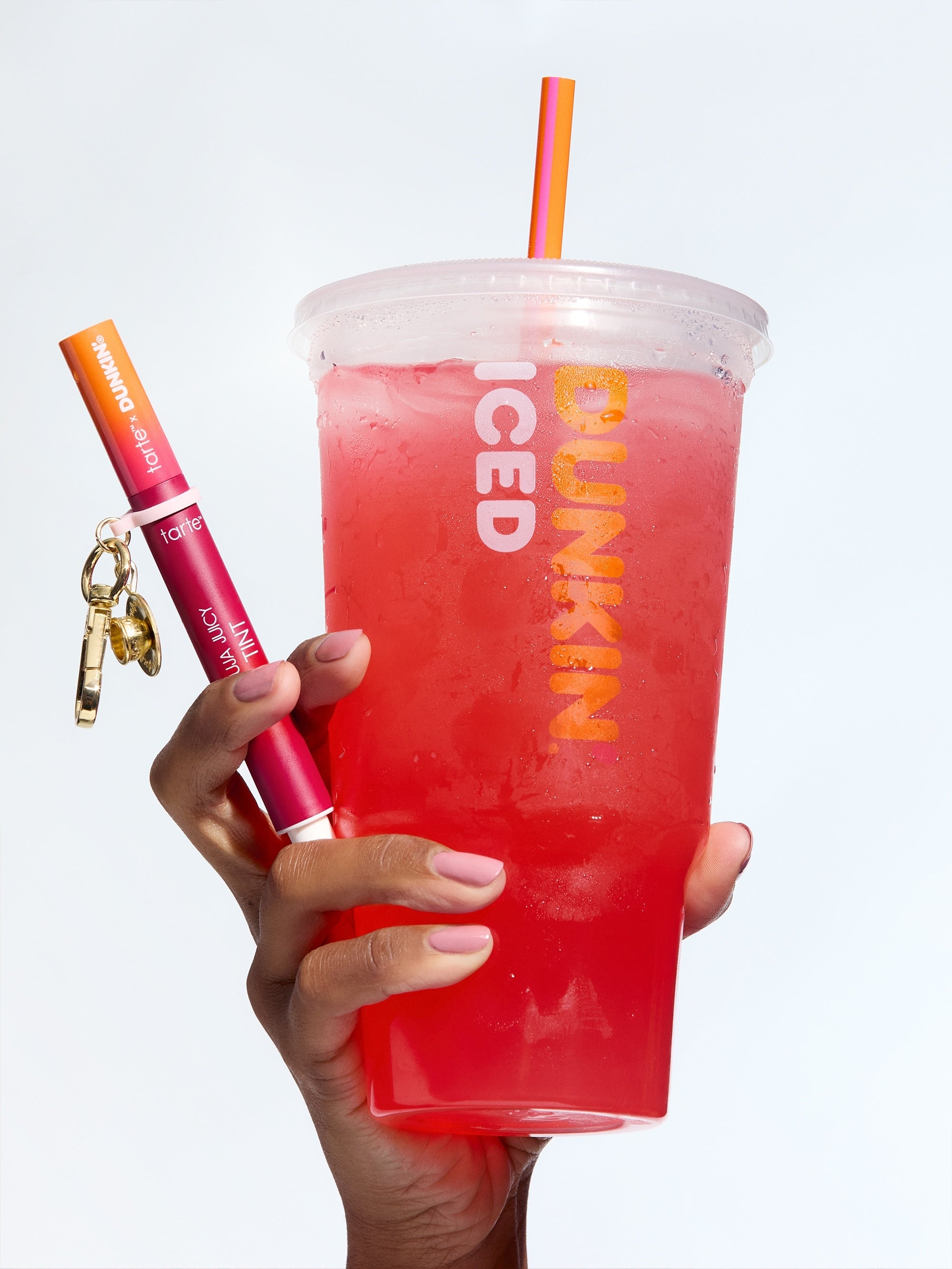 Hand holding a red iced drink from Dunkin' with a lip gloss and keychain on a white background