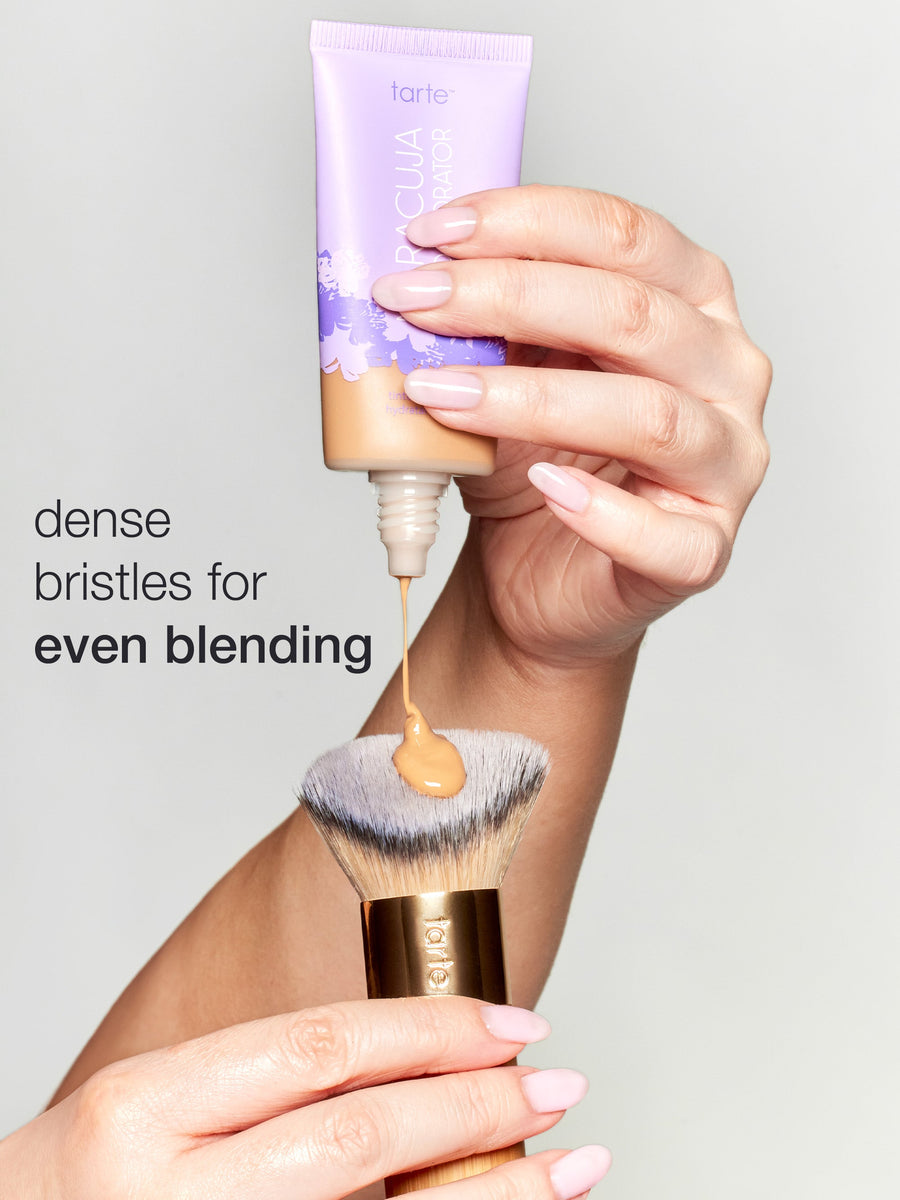 Hand holding a tarte foundation stick with a brush, against a neutral background