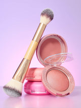 Makeup brush rests across two stacked pink blush compacts; one compact is open, revealing peach powder against a soft purple-pink gradient backdrop. Text: tarte; tarte cosmetics