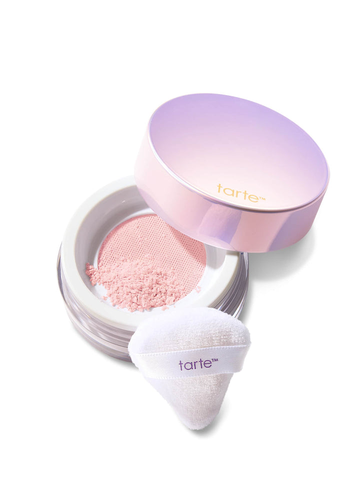 A round pastel-lavender powder jar sits open, revealing pink loose powder sifted through a mesh; a triangular white puff rests beside it on a white background. Text on lid and puff: tarte™