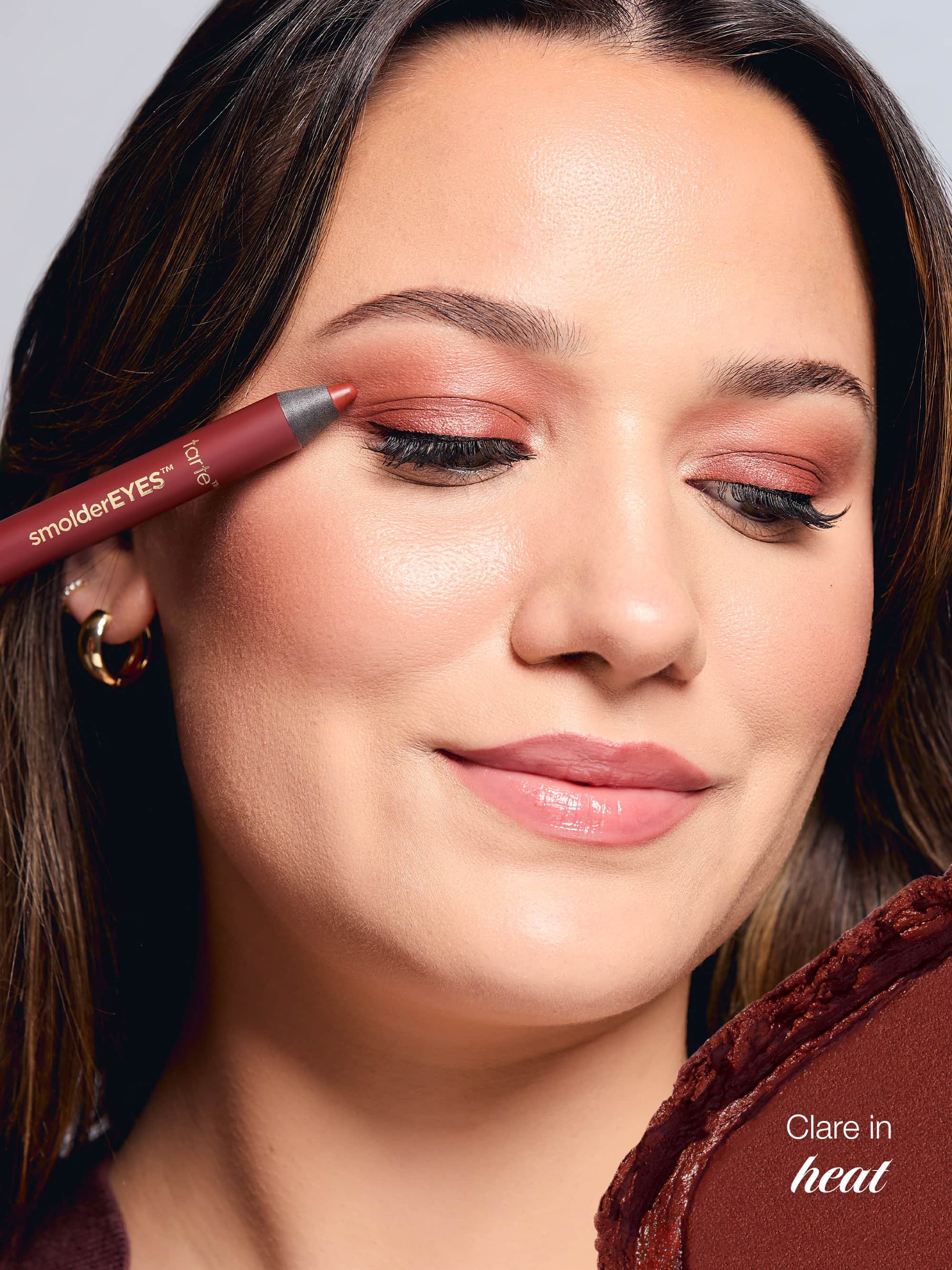 Woman applying makeup with a pencil, wearing a brown top.