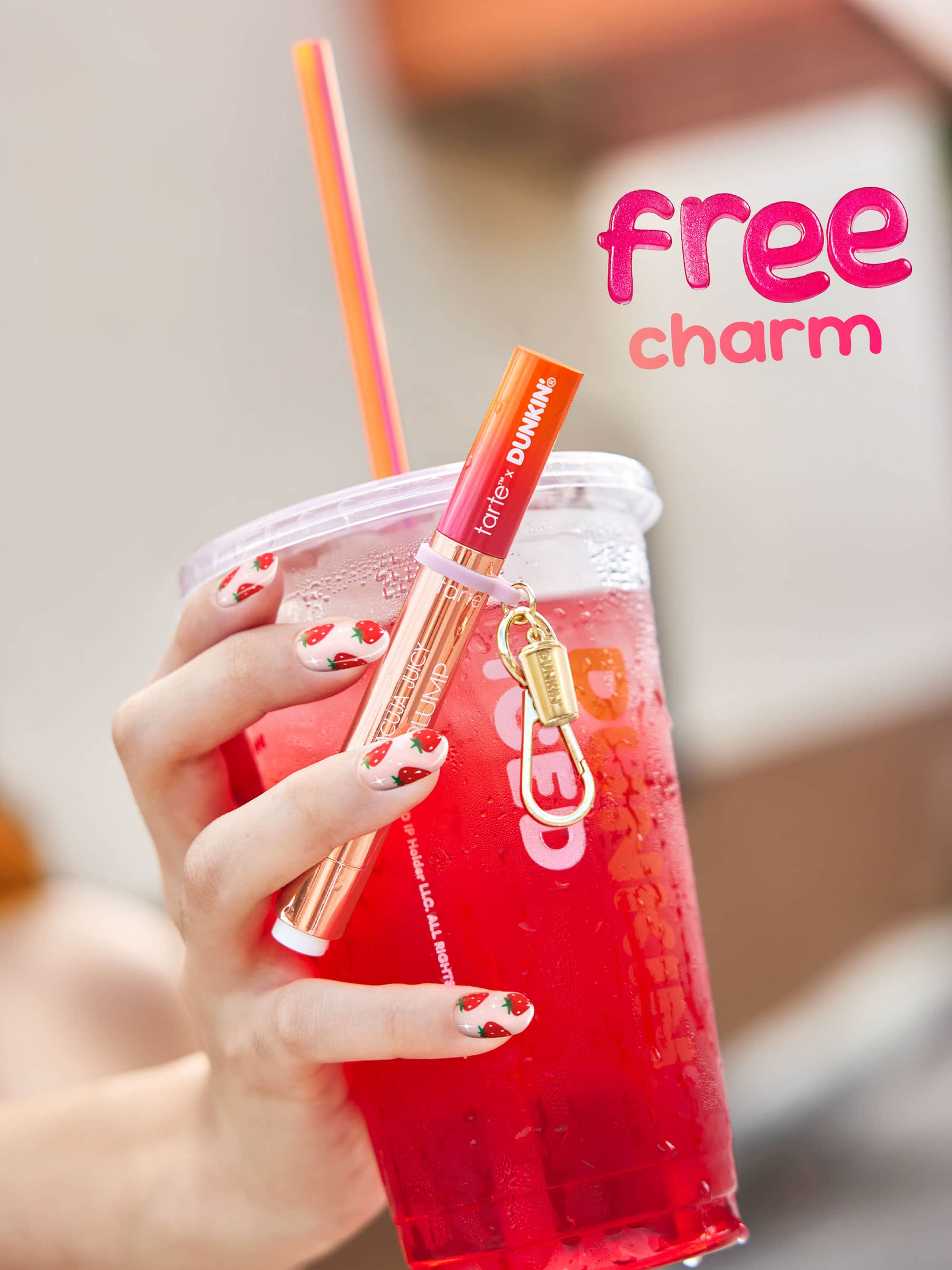 Hand holding a red drink with a straw, featuring a lip gloss charm on a blurred background.
