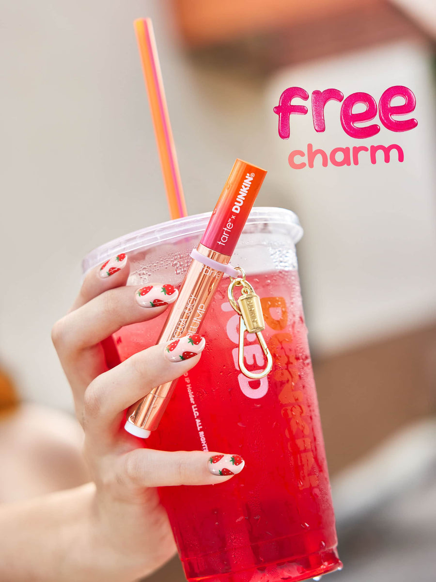 Hand holding a red drink with a straw, featuring a lip gloss charm on a blurred background.