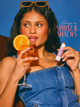 Woman holding a glass of orange juice and a lip balm with 'Spritz & Shades' and tarte maracuja juicy double ended multi stick 