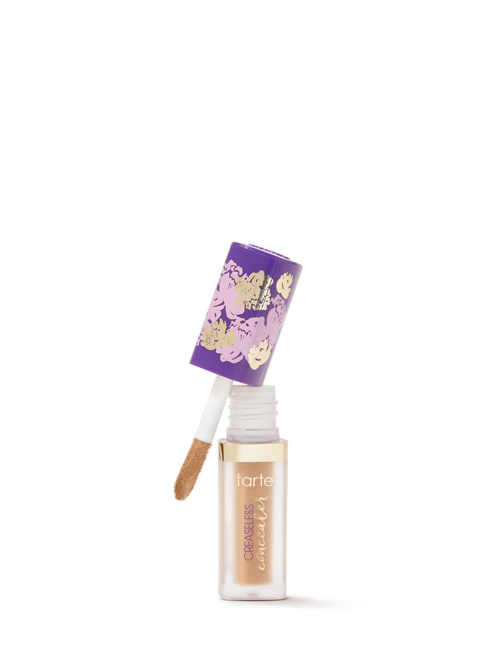 A Tarte Cosmetics concealer bottle with a beige shade features a purple floral-patterned cap, tilted and open, revealing an applicator. The background is plain white.