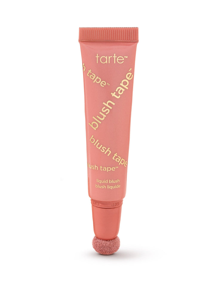A coral-pink Tarte blush tube stands upright, applicator sponge at the base, on a white background. Text visible: tarte; blush tape™ (repeated diagonally); liquid blush; blush liquide.