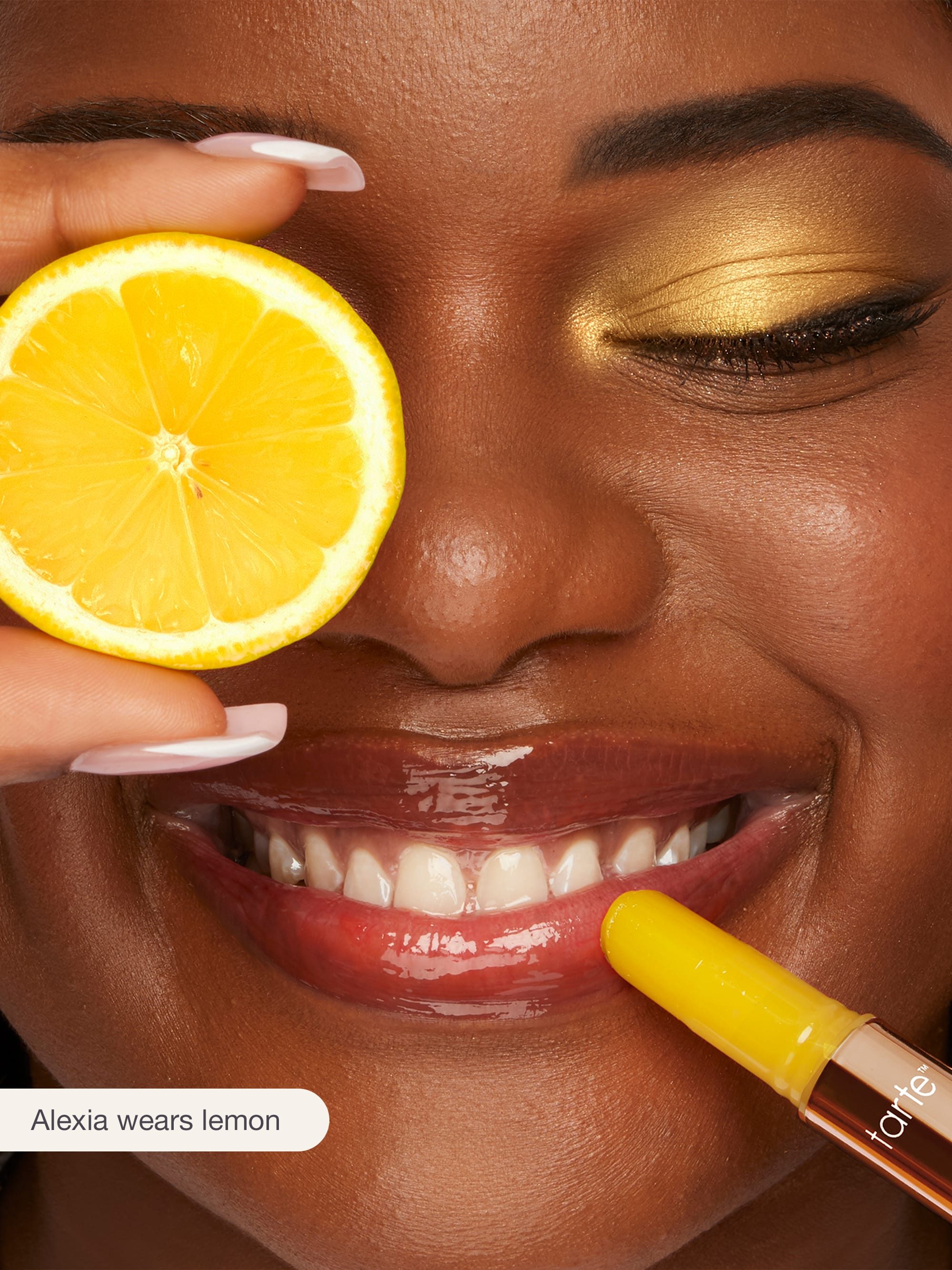 Woman holding a lemon slice to her eye with a lip gloss applicator in her mouth, wearing gold makeup.