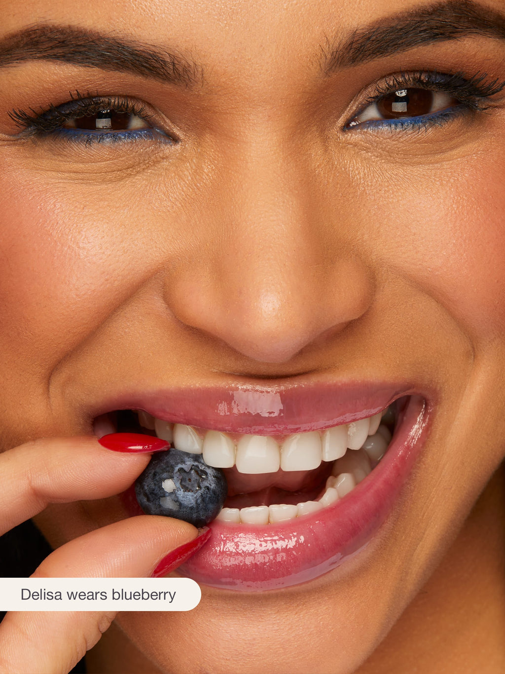 Close-up of a woman's face with blue berry in her mouth, wearing blue eyeliner.